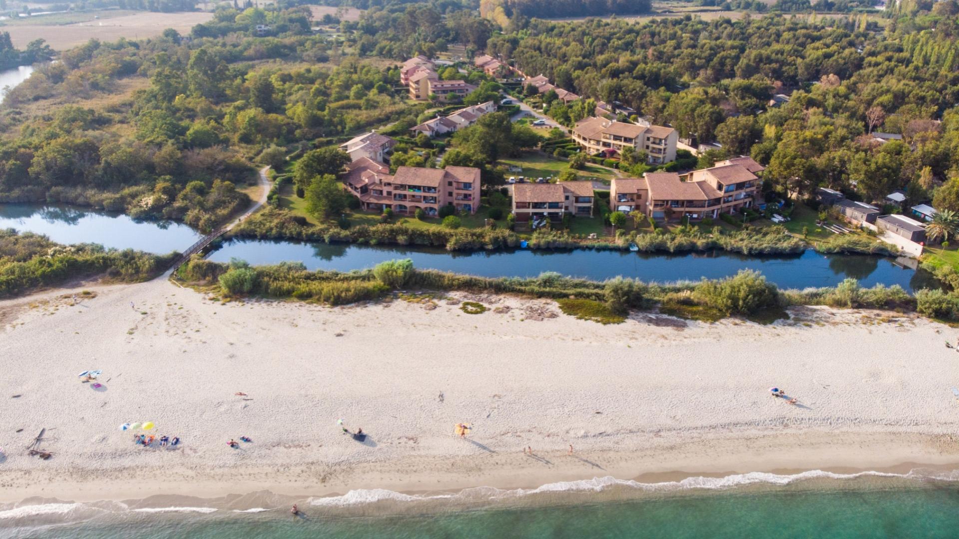 Spiaggia sabbiosa con complesso residenziale e fiume, circondati da verde lussureggiante.