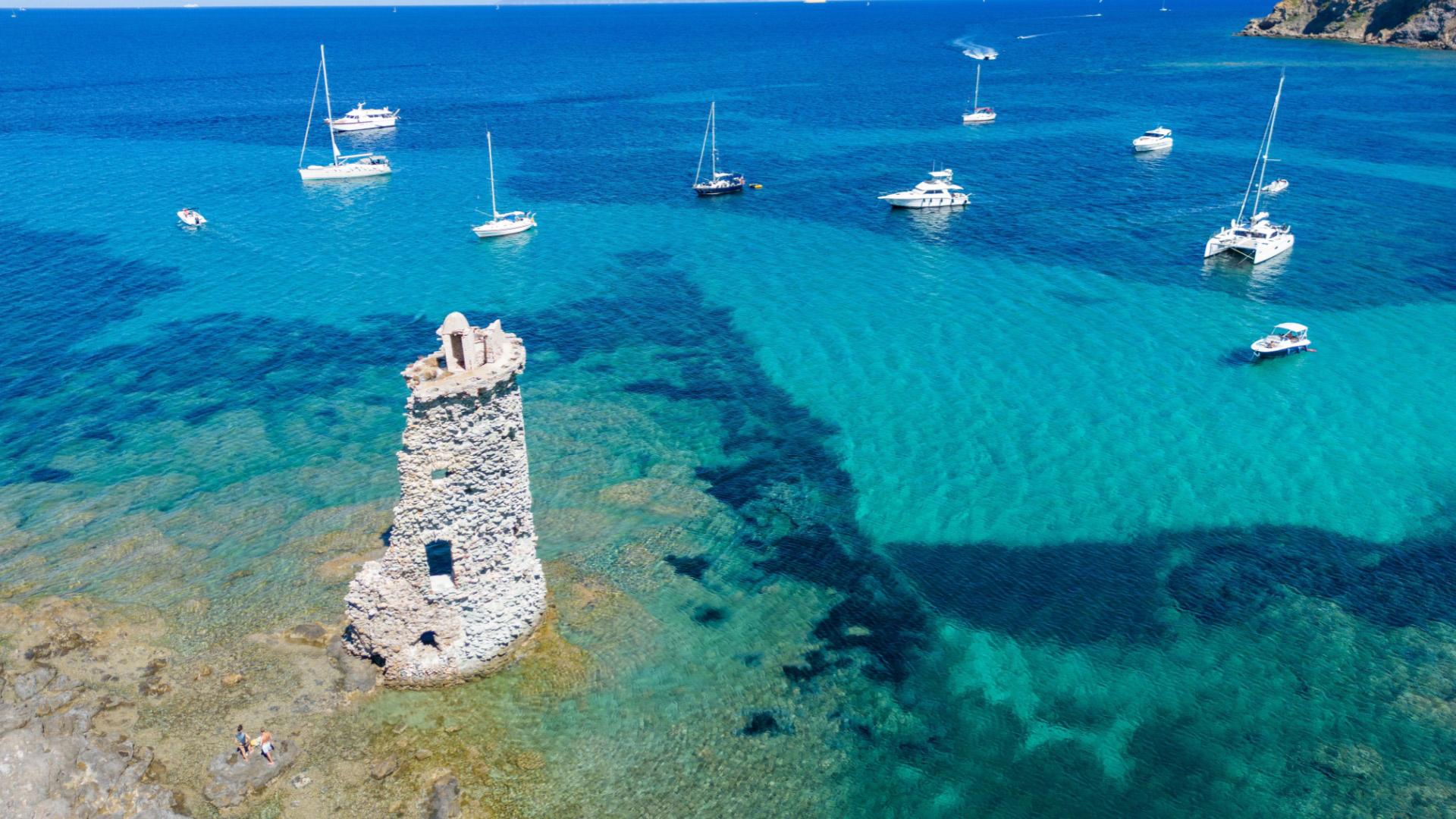 Torre costiera in pietra con barche a vela su mare turchese.