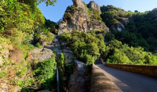 Sentiero montano con cascata e rigogliosa vegetazione lungo una strada panoramica.