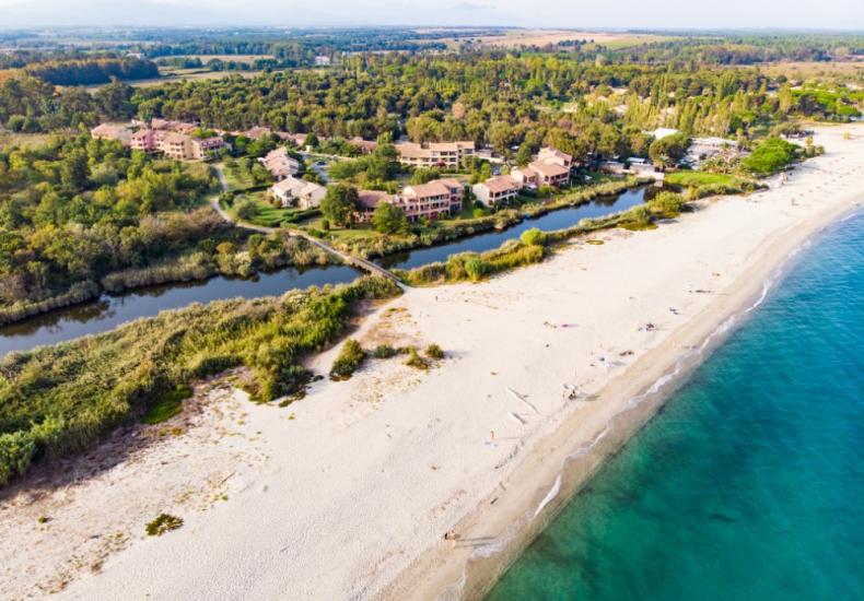 Spiaggia sabbiosa con mare cristallino e complesso residenziale immerso nel verde.