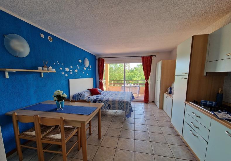 Monolocale con parete blu, letto e cucina, vista balcone.